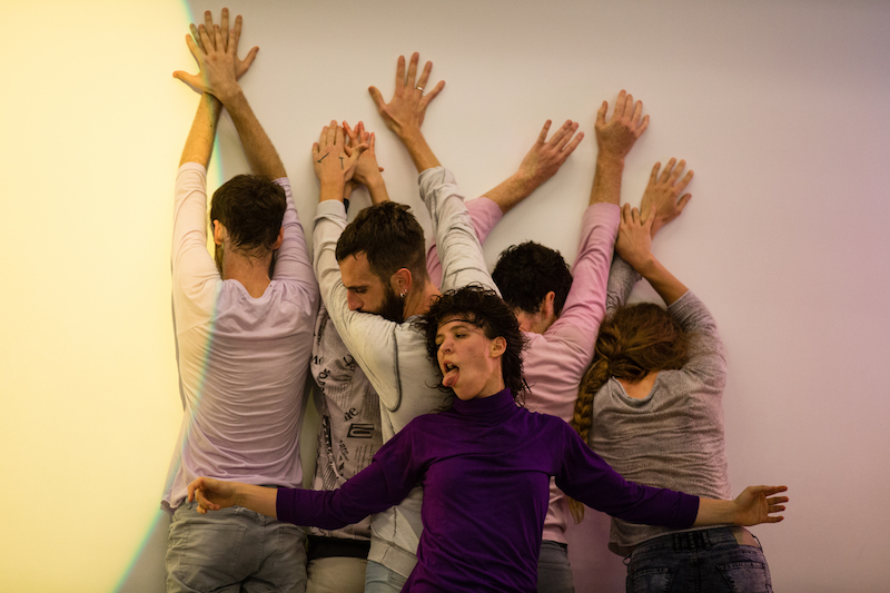A group of dancers in a clump against the wall. One woman in a purple turtleneck sticks her tongue out.